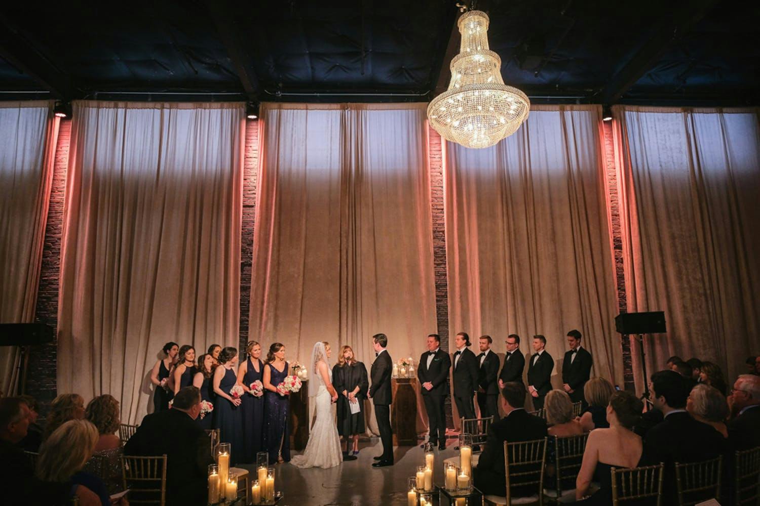 Wedding Party at Wedding Ceremony Set in Modern Industrial Loft With Exposed Brick Wall, Glittering Chandelier, and Peach Pin Spot Lighting | PartySlate