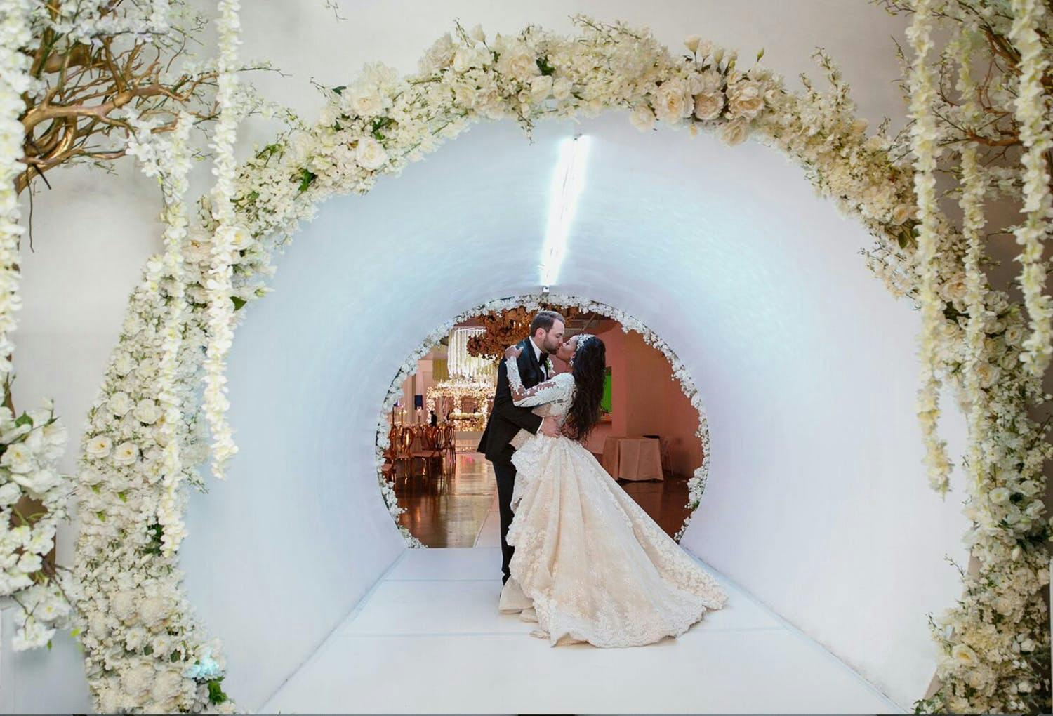 Bride and groom Kiss in Sleek White Tunnel Wedding Entrance With White Flowers | PartySlate