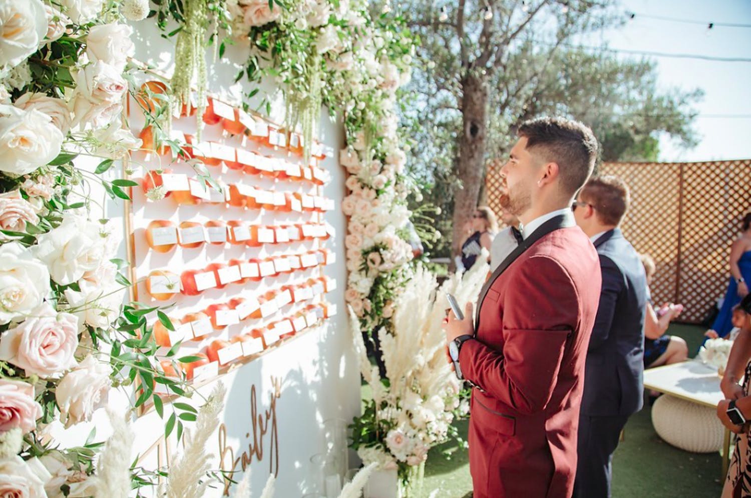 Man in Red Jacket Looks For His Wedding Seating Assignment at Floral-Framed Sign With Oranges and Place Cards | PartySlate