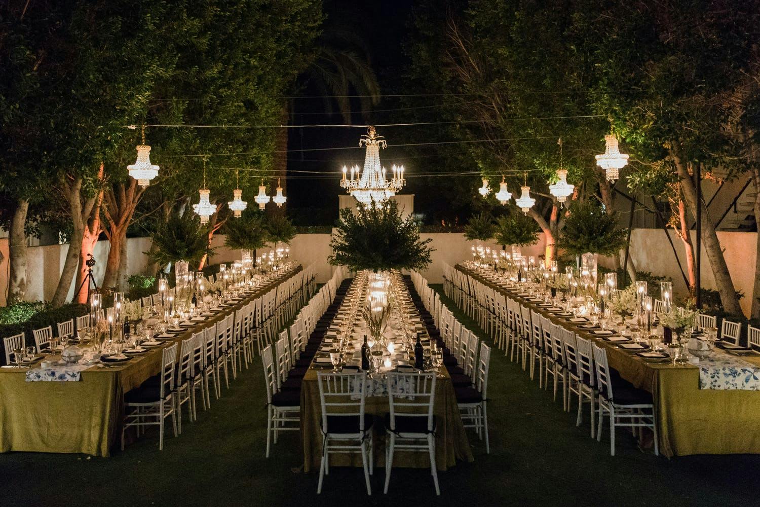 Nighttime, Outdoor Wedding Reception With Three Long Tables in Yellow Linen and Suspended Chandeliers with Greenery | PartySlate