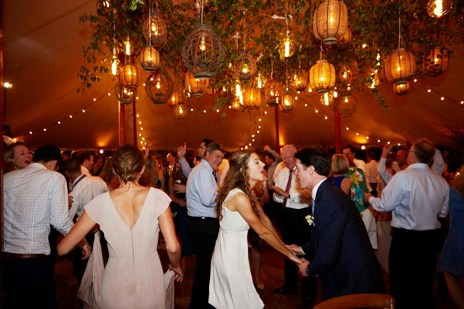 Bride, Groom, and Guests Dance in Tented Venue With Basket-Woven Lanterns and Greenery Ceiling Décor | PartySlate