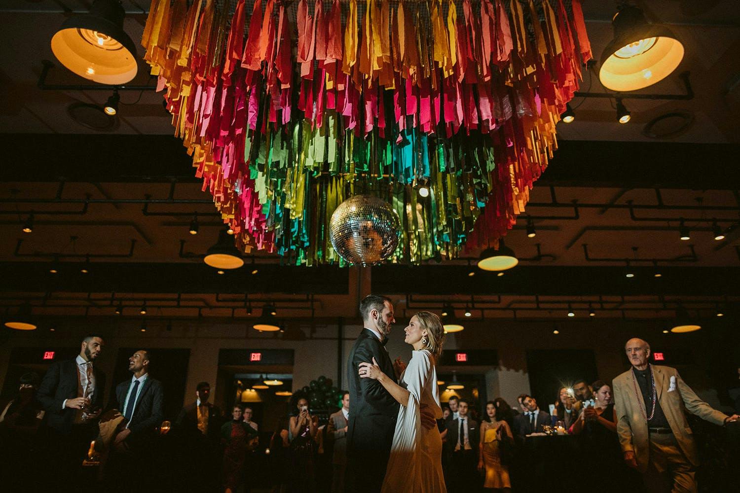 Bride and Groom Dance Beneath Wedding Ceiling Decorations of Rainbow Fringe Surrounding a Disco Ball | PartySlate