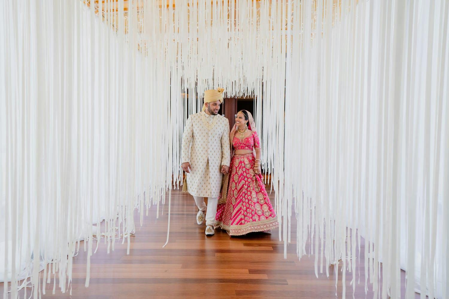 Bride and Groom Dressed in South Asian Wedding Attire Walk through White Fringe Corridor | PartySlate