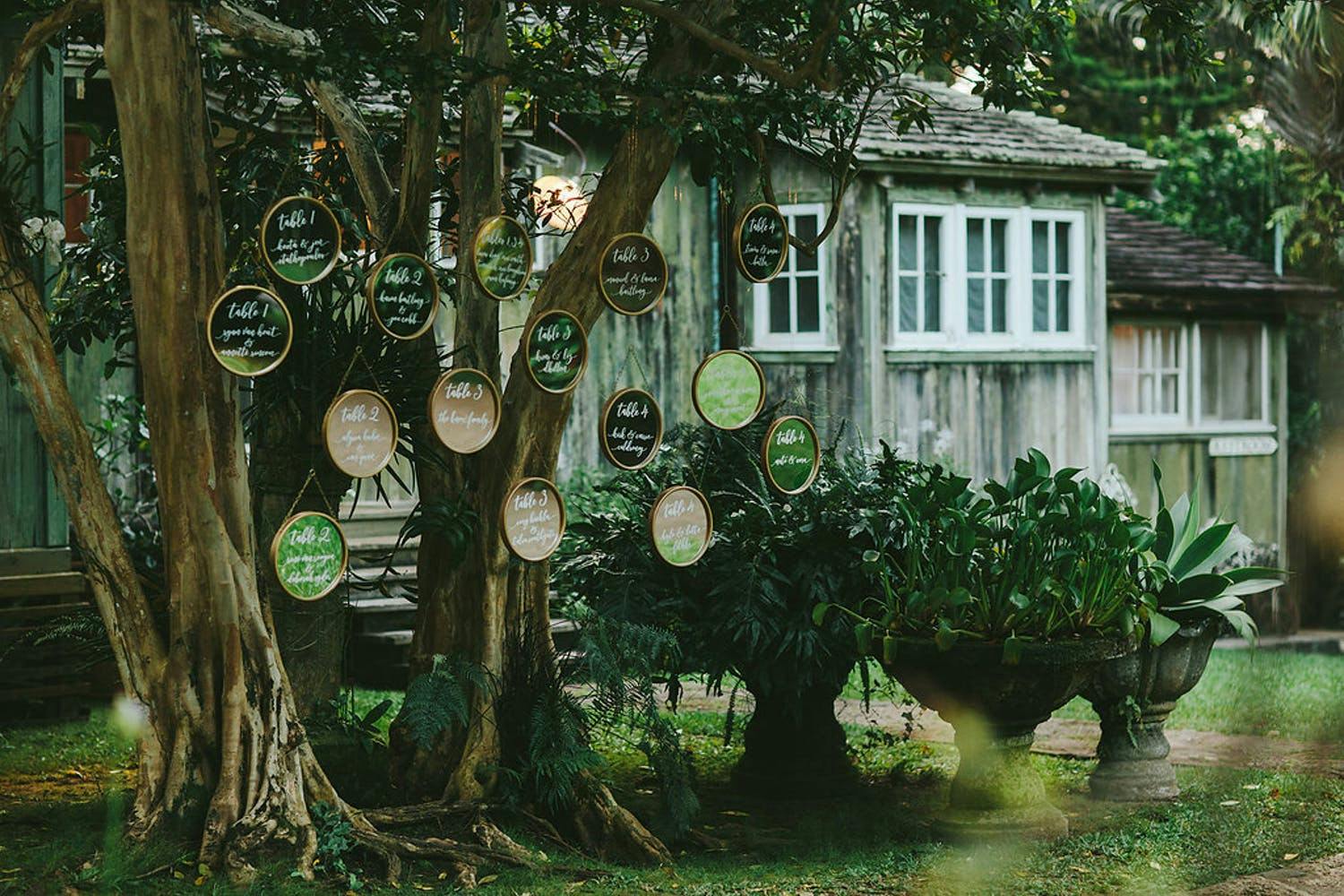 Tropical Wedding With Table Numbers Written on Green and Tan Circles Suspended from a Tree | PartySlate