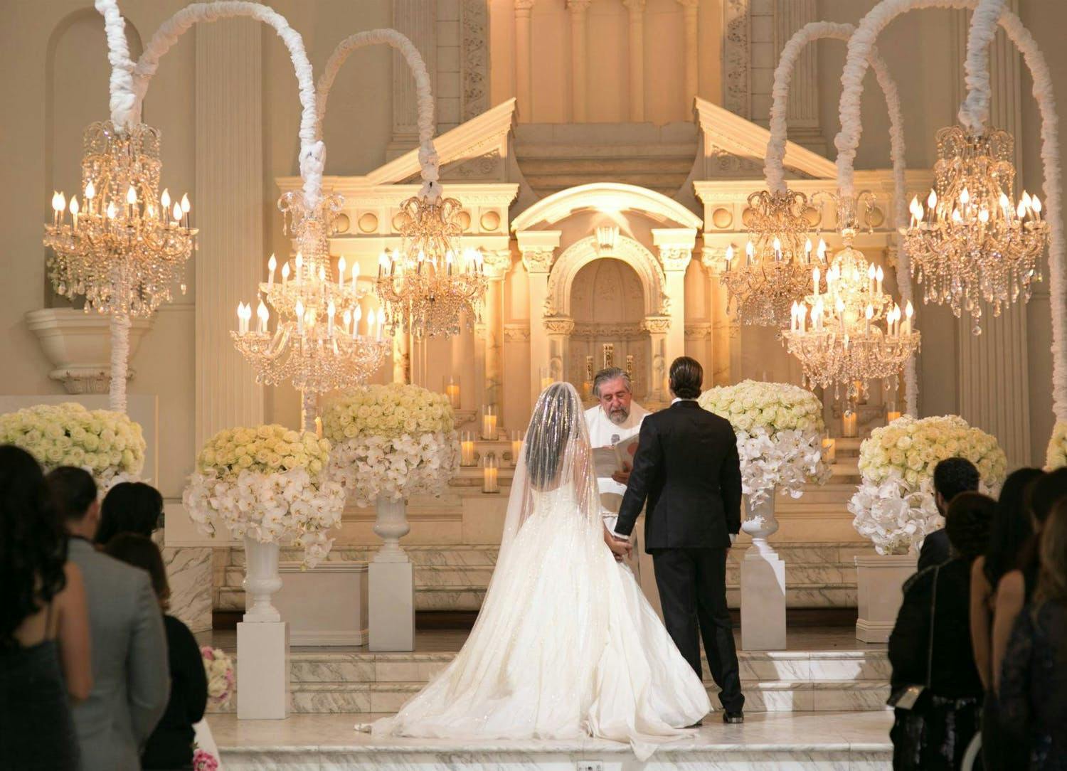 Bride and Groom Exchange Vows Before Officiant and Guests in All-White Ballroom With Chandeliers Suspended From White Curved Poles | PartySlate
