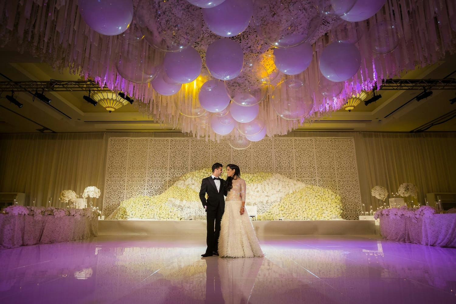 Жених и невеста стоят на танцполе с фиолетовой подсветкой и под свадебным потолком, украшенным мерцающими воздушными шарами | PartySlate