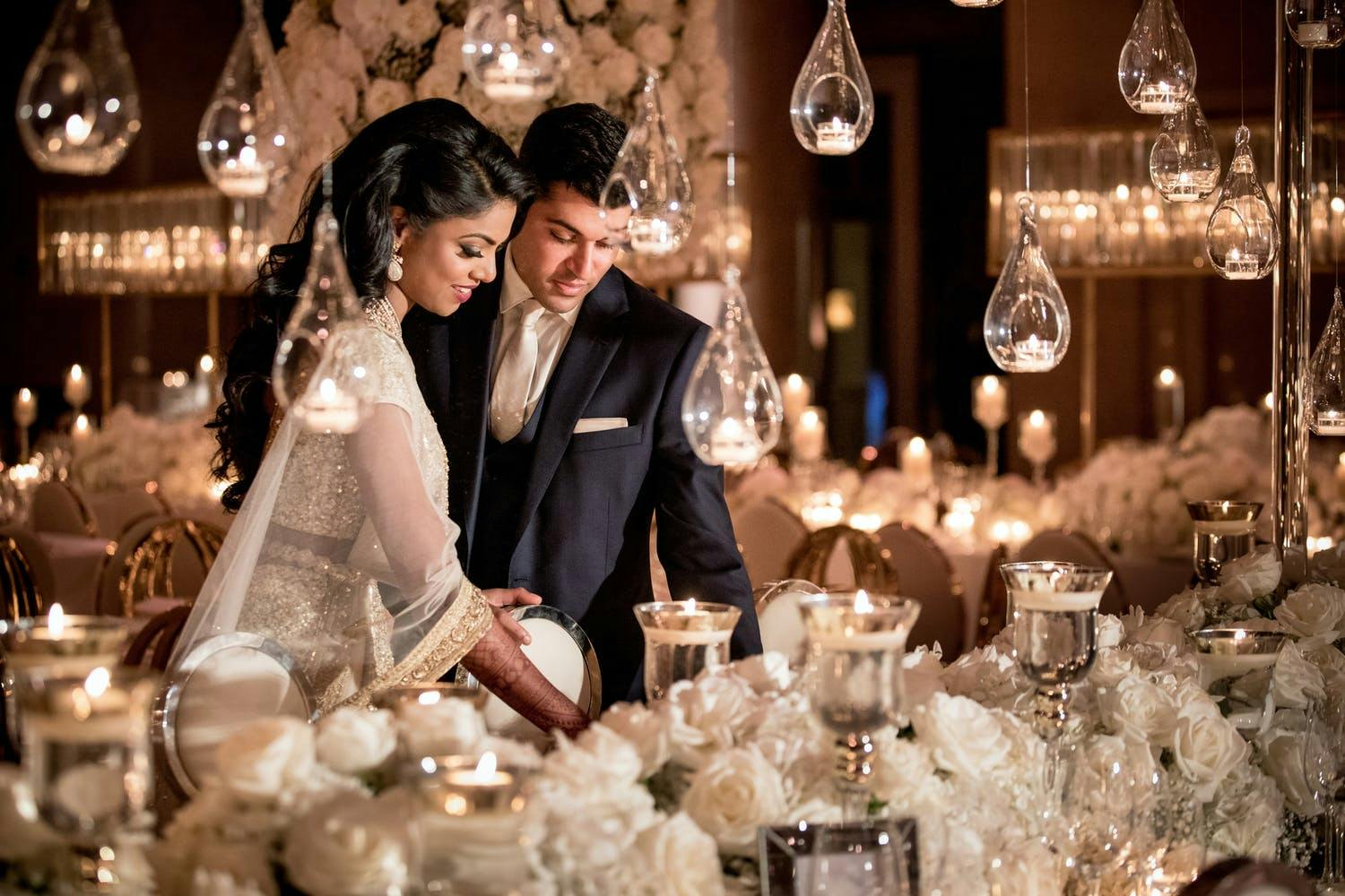 Bride and Groom Share a Moment at White Wedding Tablescape and Suspended Globed Candlelight | PartySlate