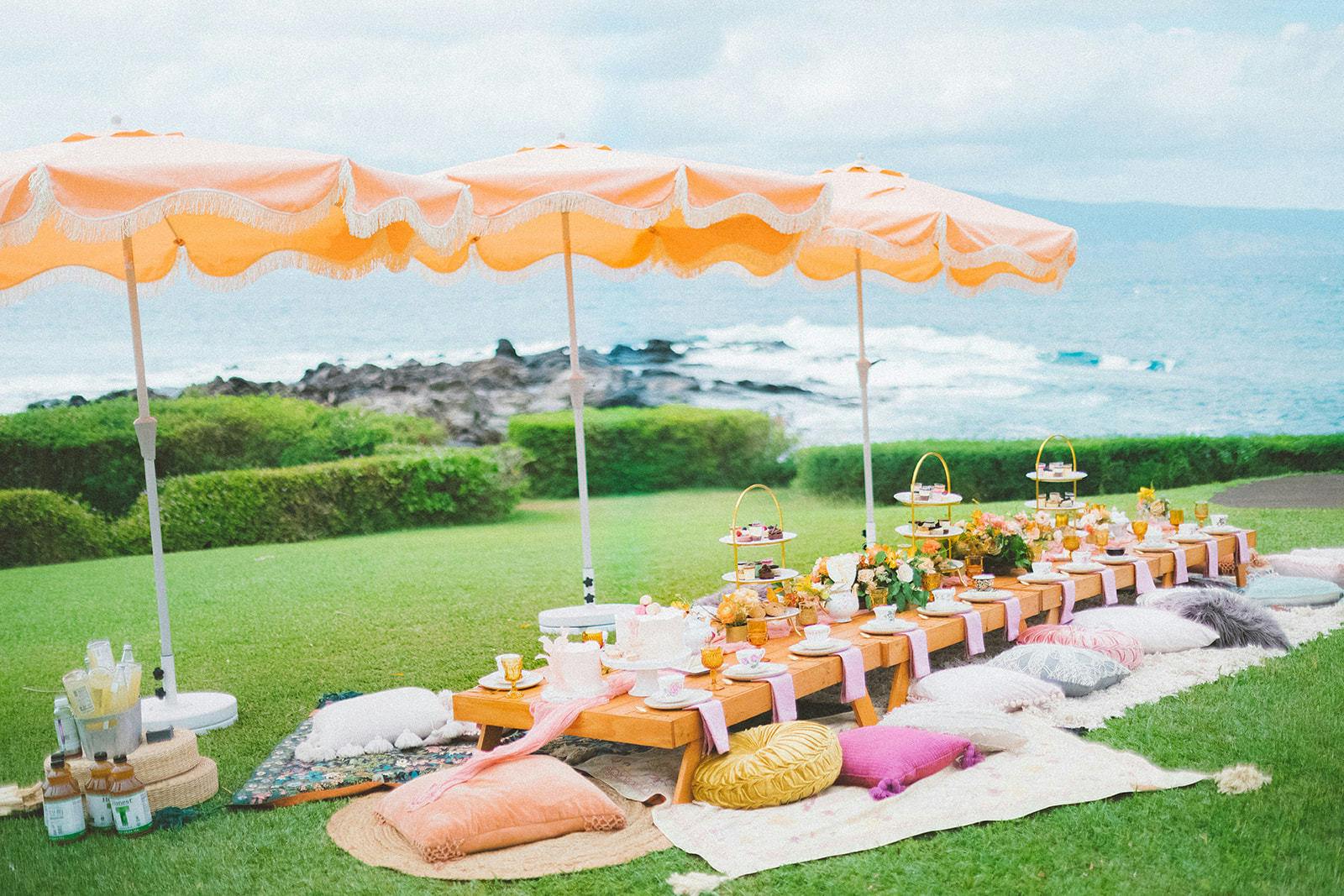 Tropical Orange Pink and White Oceanfront High Tea Party at Montage Kapalua Bay in Kapalua, HI with Ocean in the Background and Pillows on the Ground Around a Low Table with Umbrellas | PartySlate