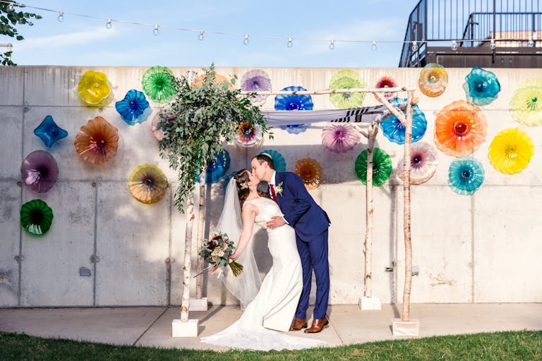Bride and Groom Kiss Beneath Chuppah in Funky and Colorful Urban Garden at Ignite Glass Studios in Chicago, IL | PartySlate