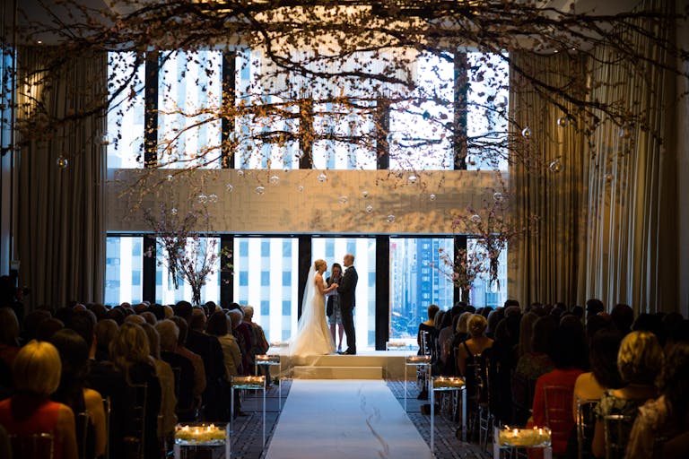 Bride and Groom Exchange Vows Indoors at The Langham, Chicago With Floor-to-Ceiling Windows and Overarching Branches | PartySlate