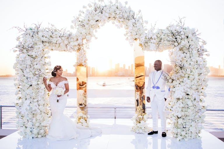 Bride and Groom Pose in Front of a Mirrored Trifold Gold Arch Covered in White Flowers | PartySlate