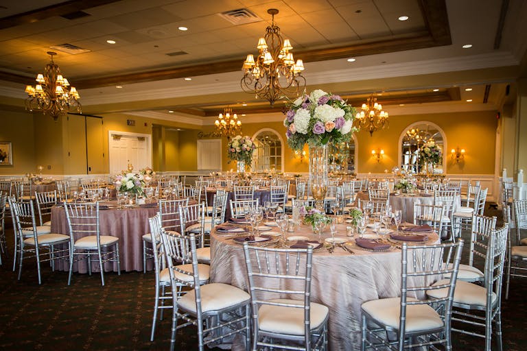 Reception room filled with tables with purple floral centerpieces and stunning gold chandeliers at White Eagle Golf Club in Naperville, IL | PartySlate