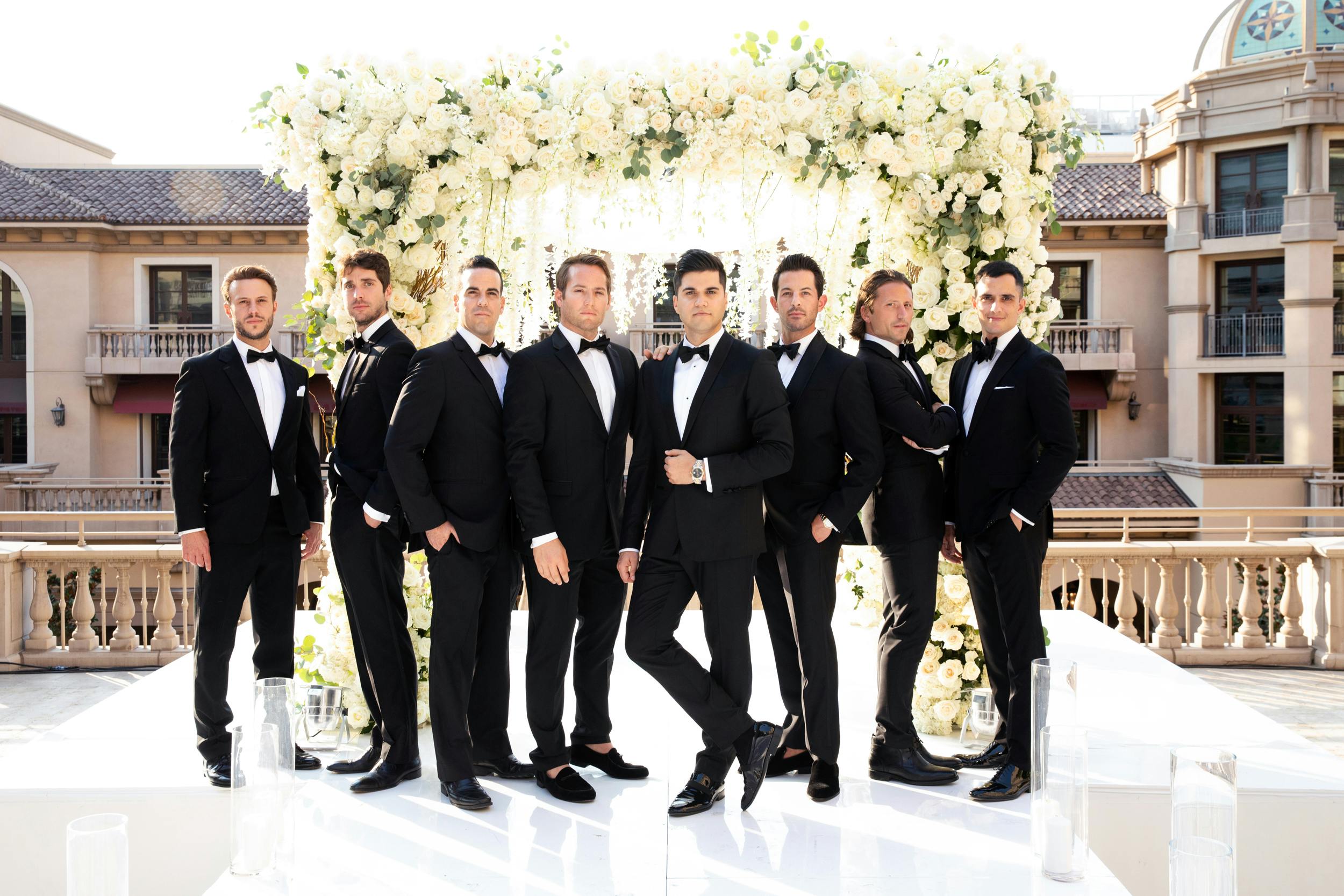 8 groomsmen in black tuxedos striking a pose in front of wedding arch | PartySlate