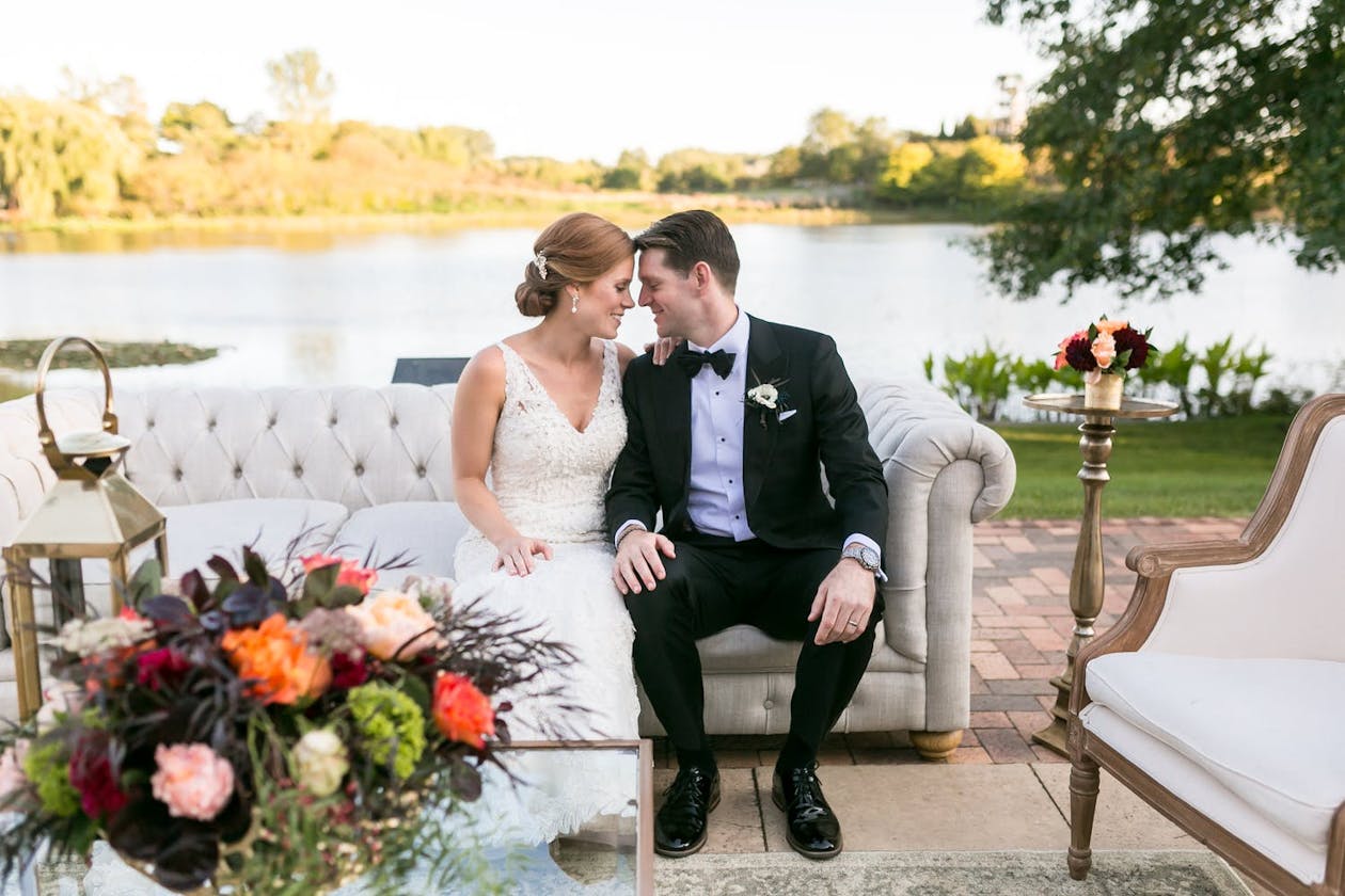 Bride and Groom Sit on White Sofa Against Lake Backdrop at Chicago Botanic Garden in Glencoe, IL | PartySlate