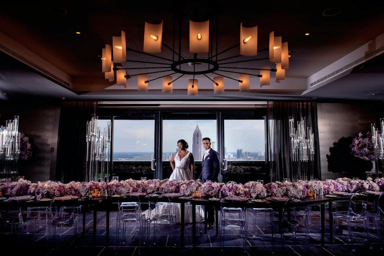 Bride and Groom Stand Between Pink Floral-Lined Tablescape and Windows with City Views at Four Seasons Hotel Atlanta in Atlanta, GA | PartySlate