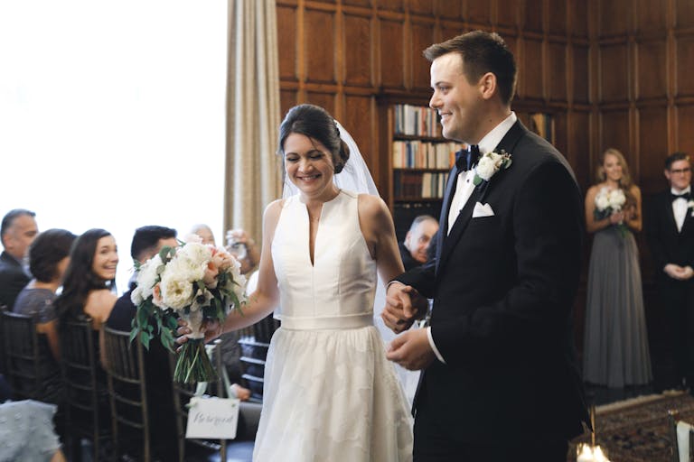 Bride at groom walk down aisle with shelved books in backdrop at The Quadrangle Club in Chicago, IL | PartySlate