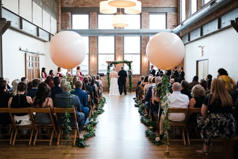 high ceiling Art Deco space with greenery lined wedding aisle with two giant white balloons | PartySlate