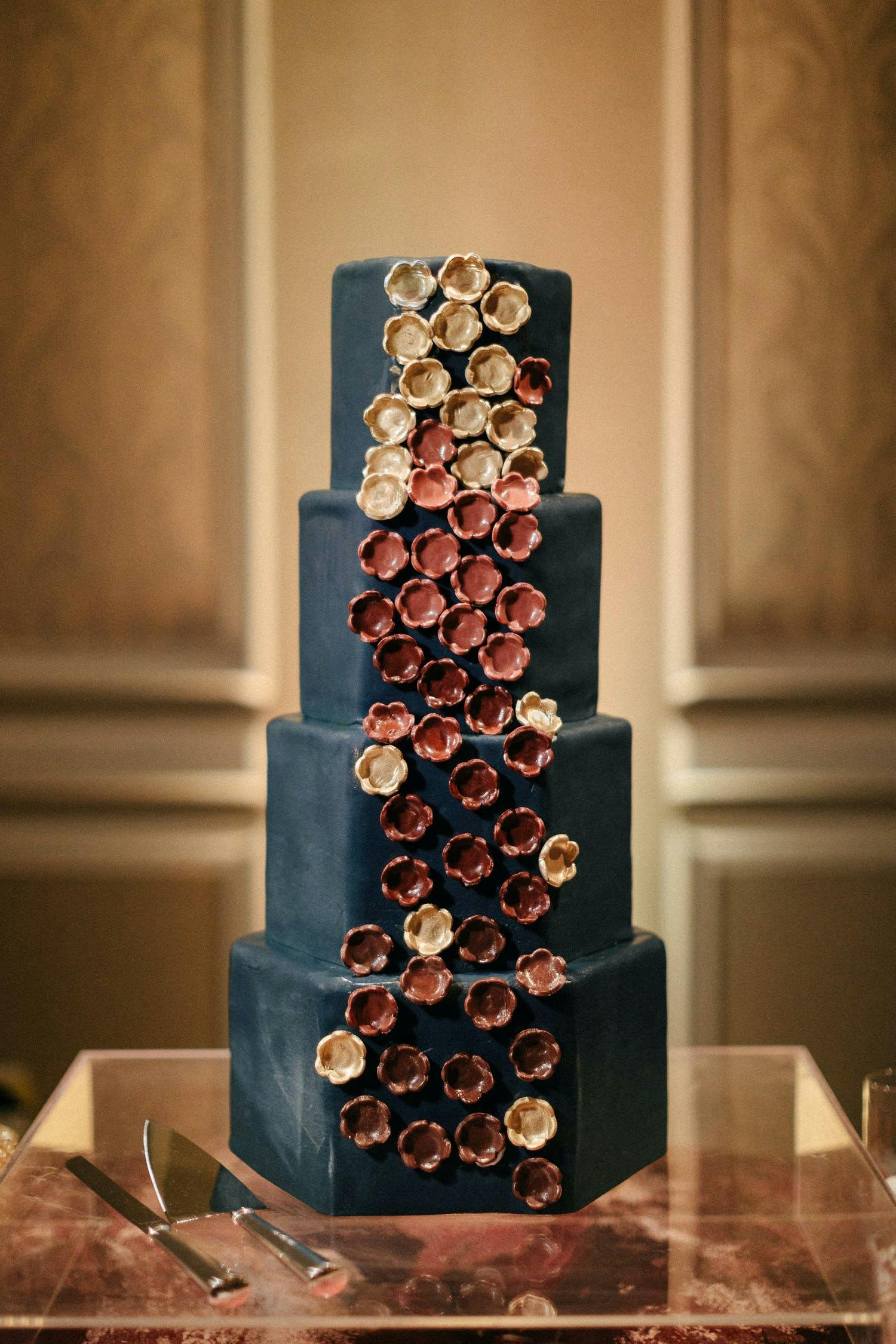 Four-tier black wedding cake with burnt-red, pink, and gold candied flower designs down side | PartySlate