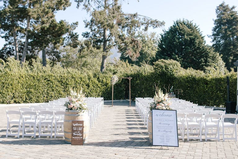 Outdoor wedding ceremony with white chairs and floral-decorated barrels bordered by manicured trees at Folktale Winery & Vineyards in Carmel-by-the-Sea, CA | PartySlate