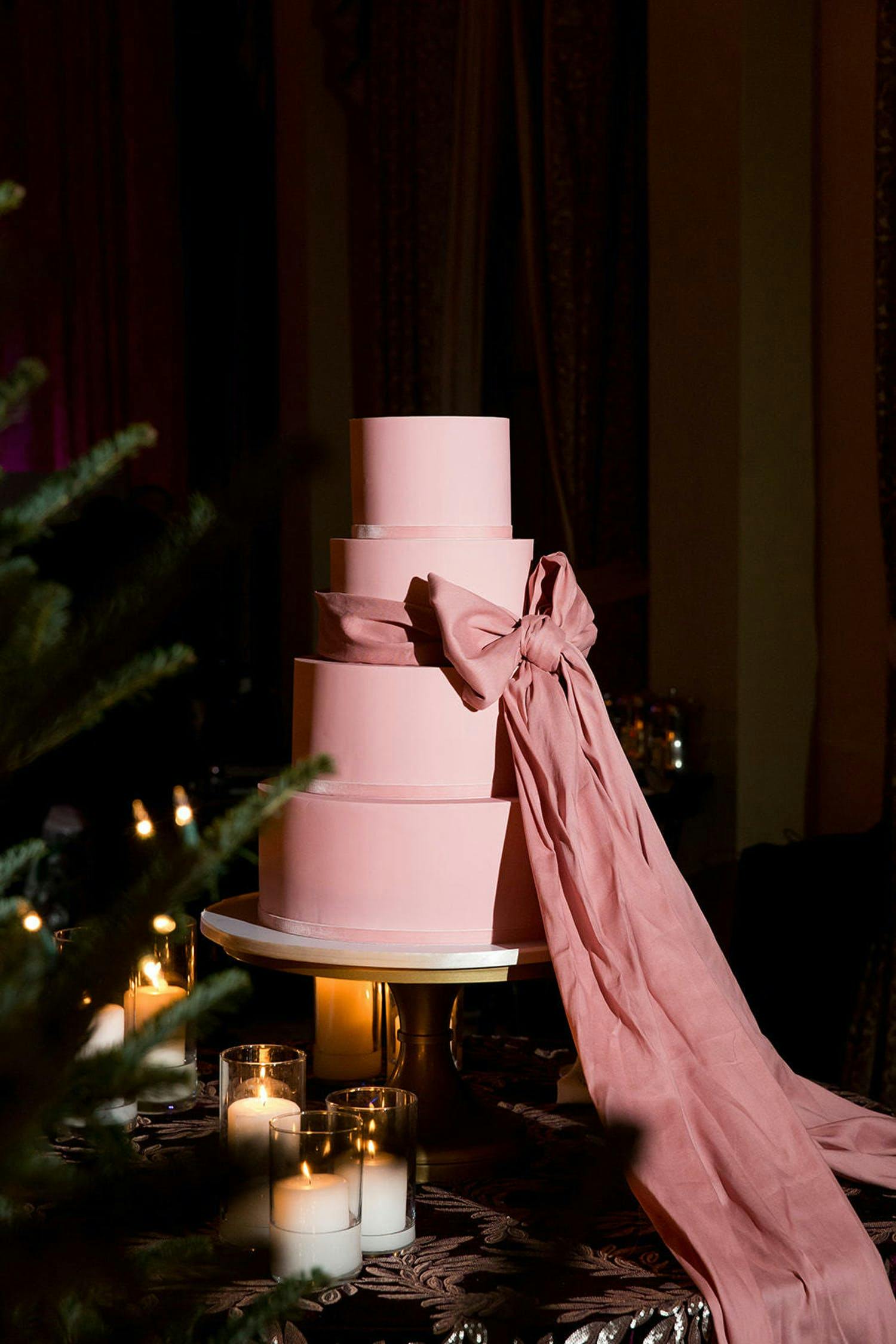 Four tier light pink wedding cake with big pink bow wrapped around it | PartySlate