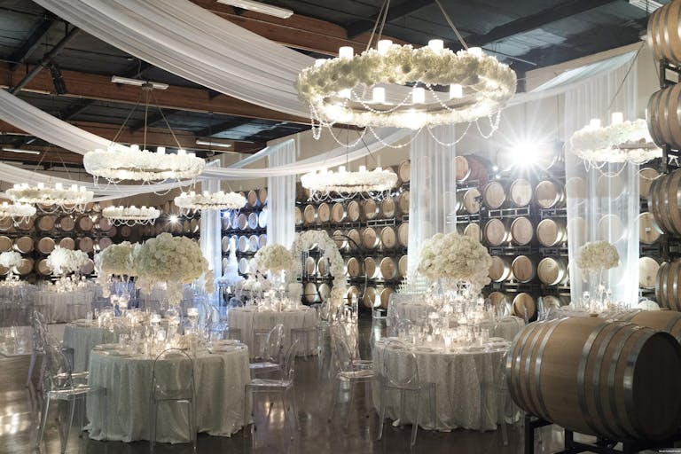 White tablecloths, floral centerpieces, and draping tapestries at white themed wedding in Callaway Vineyard and Winery’s barrel room | PartySlate
