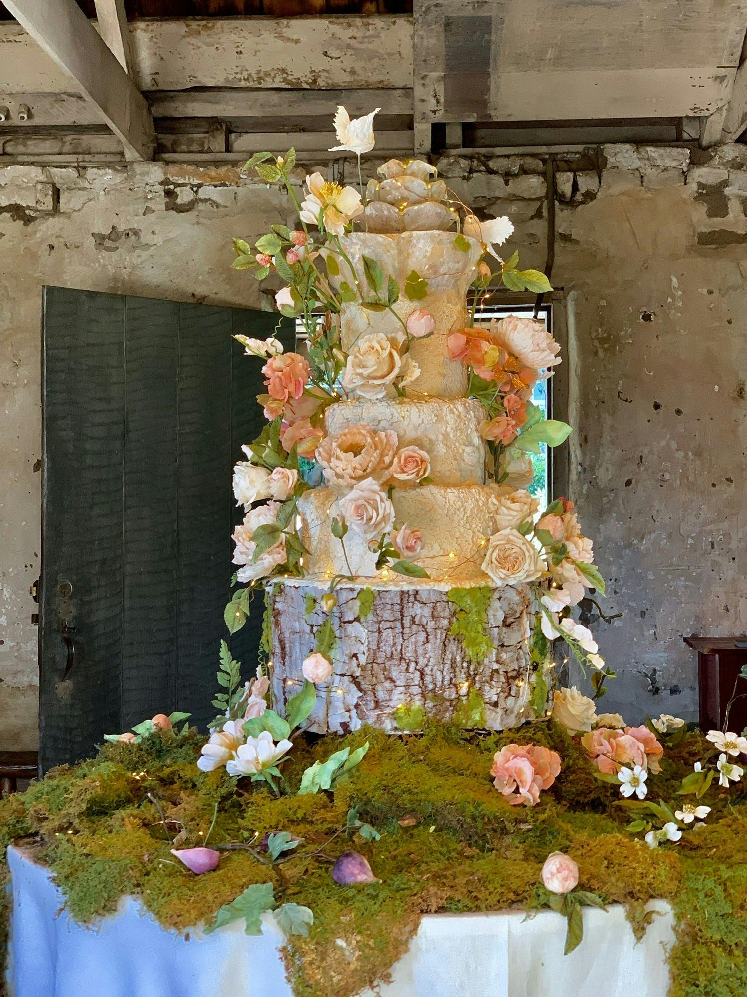 Large wedding cake on tree-trunk base and bed of moss with tiers covered in nature-like floral designs | PartySlate