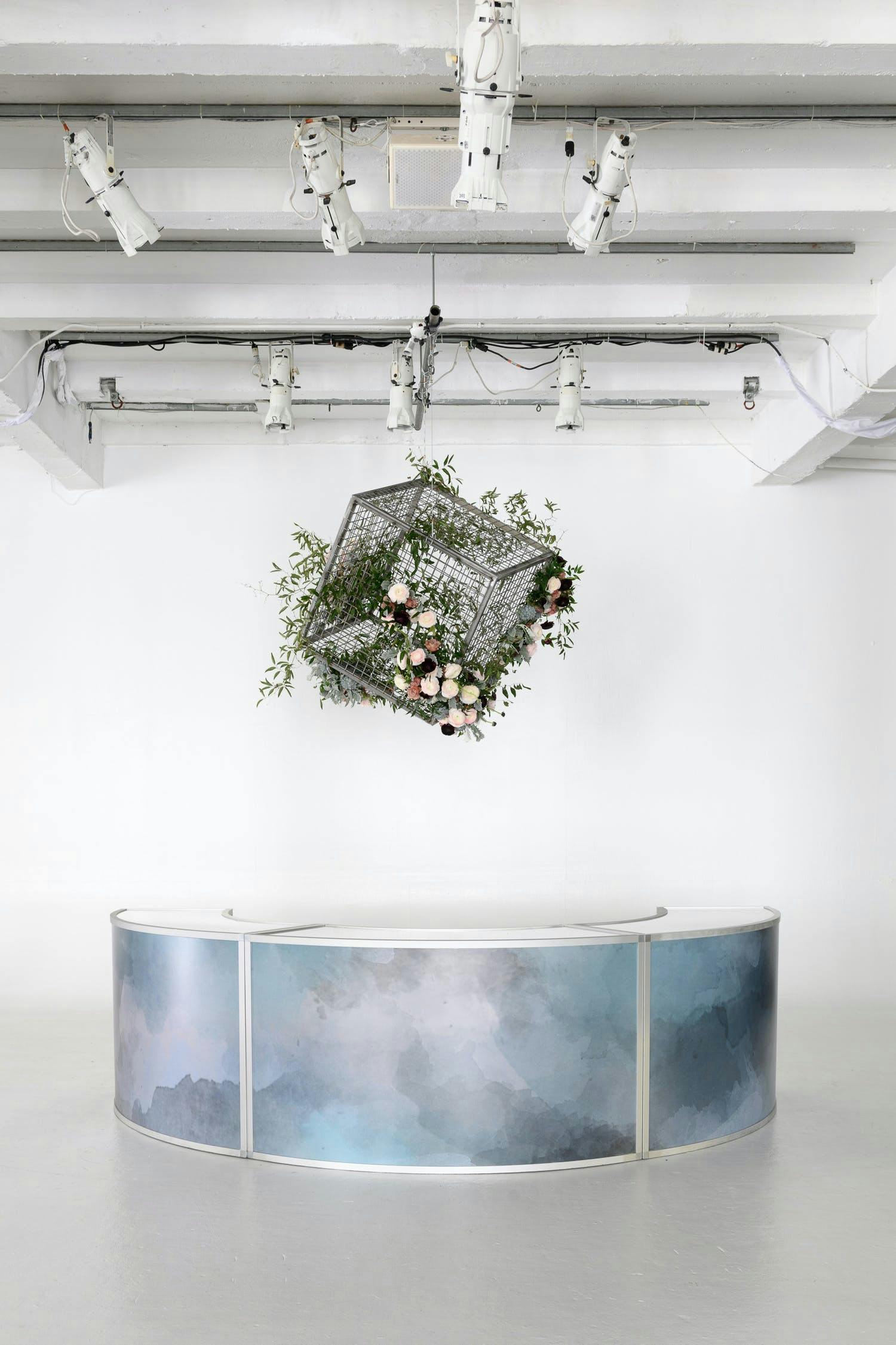 A white raw space with a galaxy patterned reception table against a white wall.. A black cube cage hangs above with greenery and pink florals | PartySlate