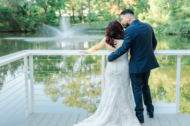 Groom kisses bride's head as she looks at pond at Forever 5 Events in Conroe, TX | PartySlate