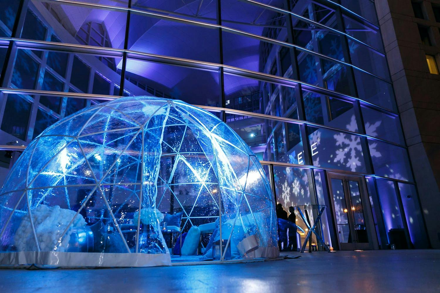 Transparent glamping igloo is lit up by blue lights in front of a wall of windows at winter blue corporate event party | PartySlate