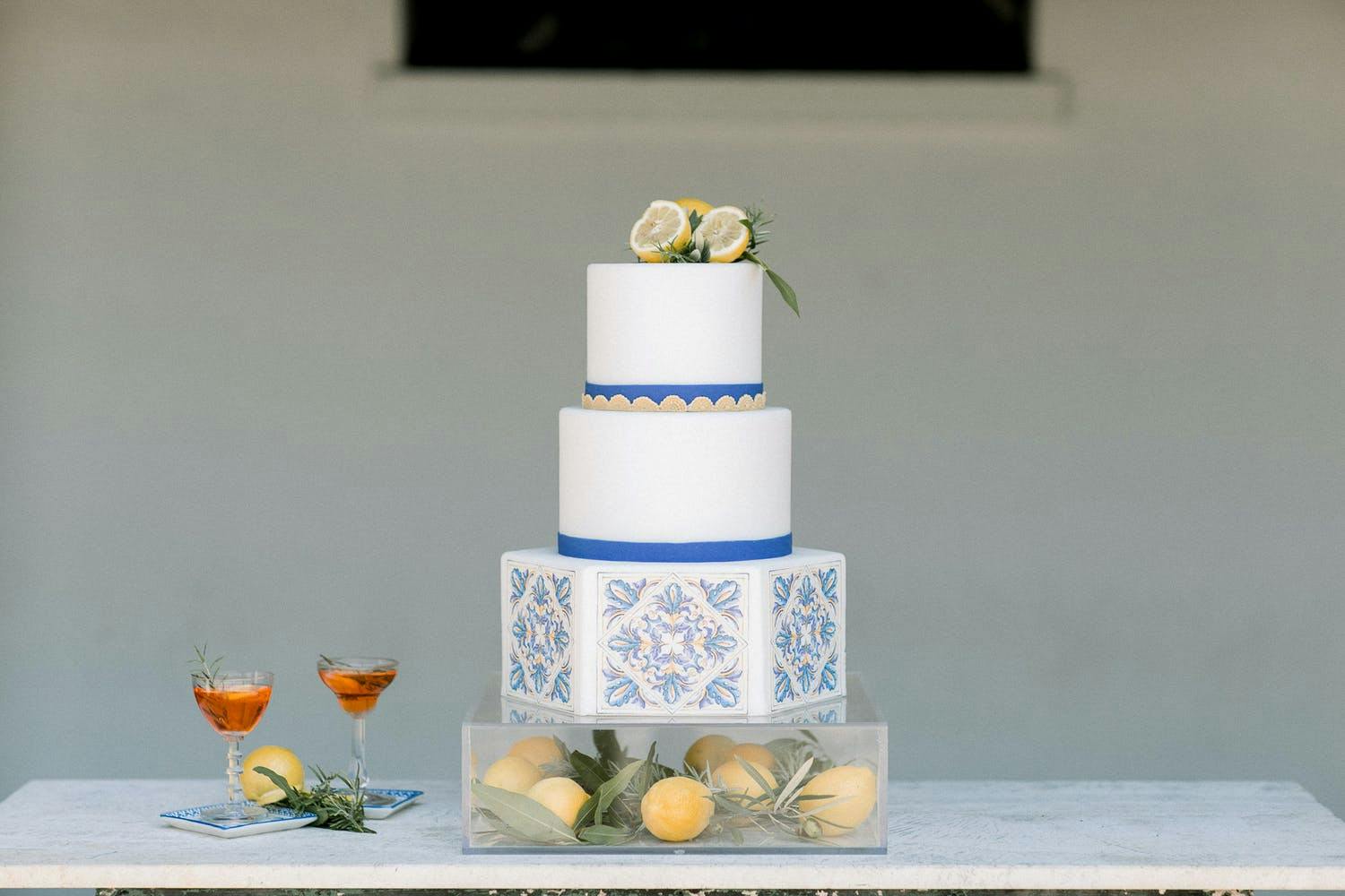 Three-tier wedding cake with a hexagon bottom tier and blue and white design on a lucite cake stand filled with lemons | PartySlate