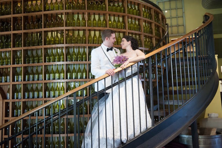 Bride and groom smile at each other on stairway curving around towering wine wall at City Winery Chicago in Chicago, IL | PartySlate