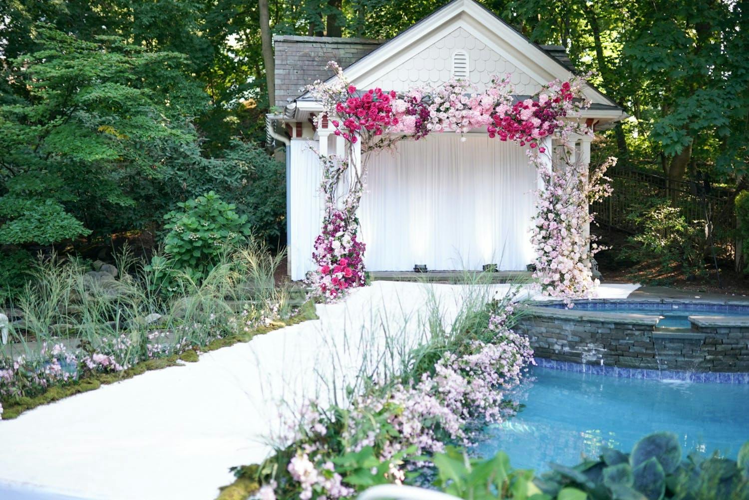 Backyard wedding with white aisle running alongside inground pool and pink floral arch against small white building | PartySlate