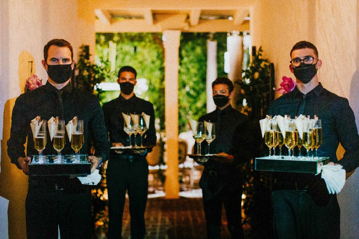 Four male servers in black wear face masks and gloves and carry trays of Champagne for Event During COVID-19 | PartySlate