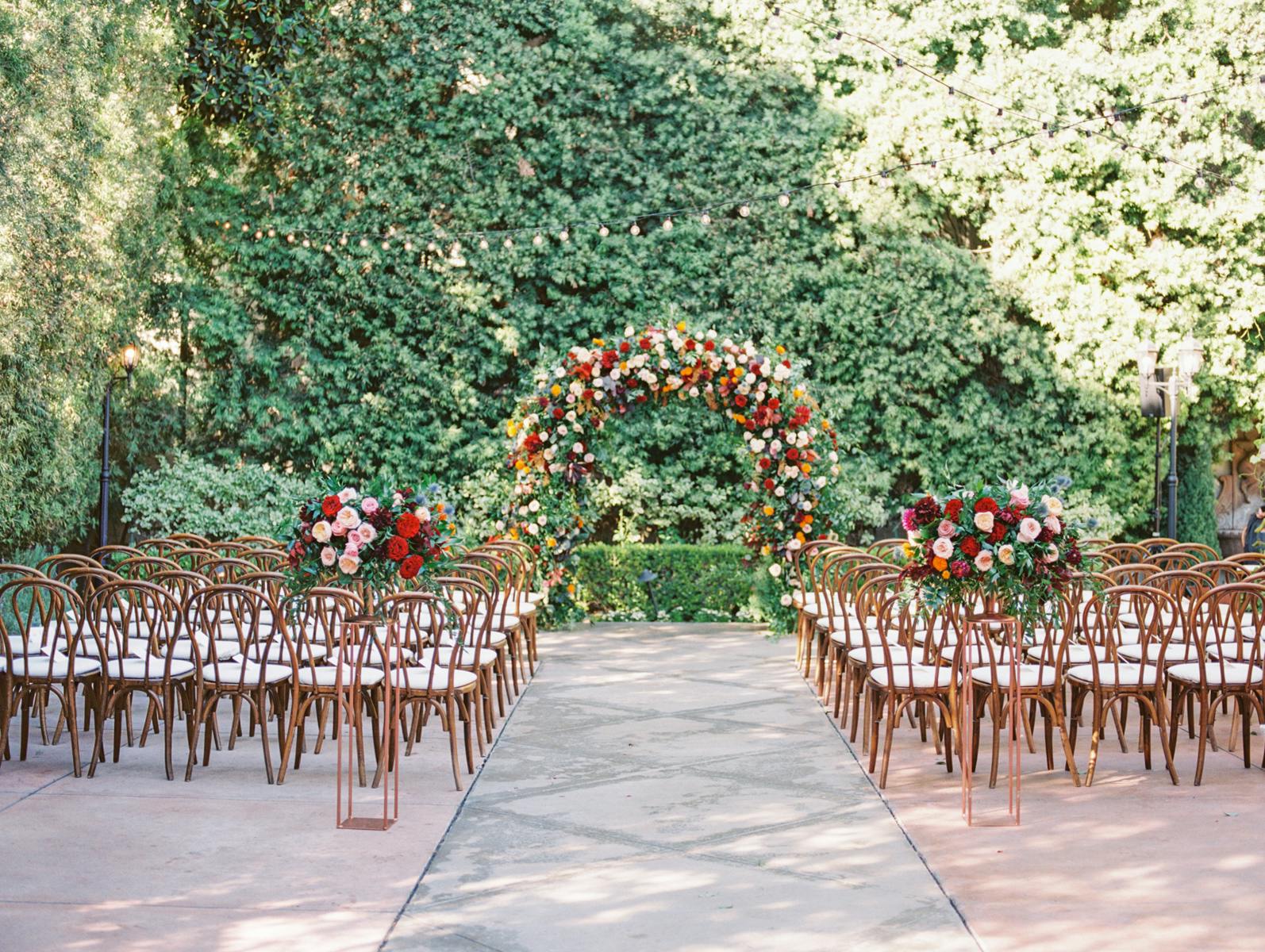 An outdoor wedding ceremony with fall wedding decorations that include a red and green floral arch | PartySlate