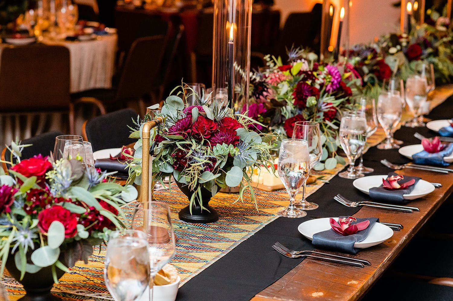 Wedding reception table with crimson and greenery centerpieces in jet-black vases | PartySlate