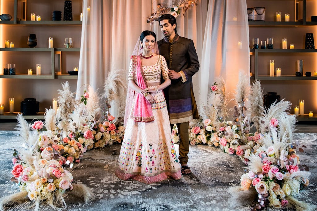 Bride and groom stand in front of dreamy pink drapery and half circle of papas grass and pink blooms at South Asian wedding | PartySlate