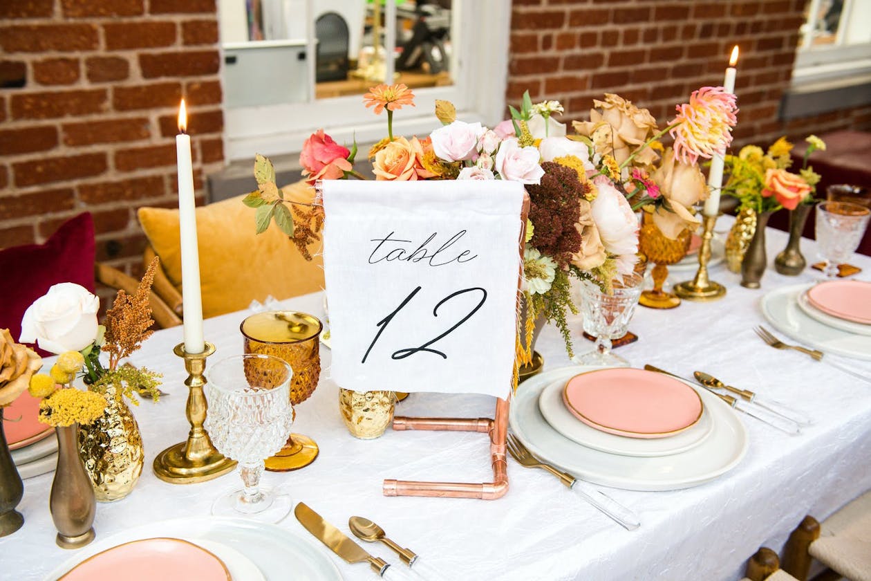 An all-white table setting with pink accented plates, a white linen table number, and bright floral centerpieces | PartySlate