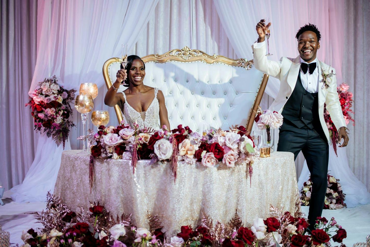 Bride and groom toast at sweetheart table with pink and red flowers on table and at foot of the table | PartySlate