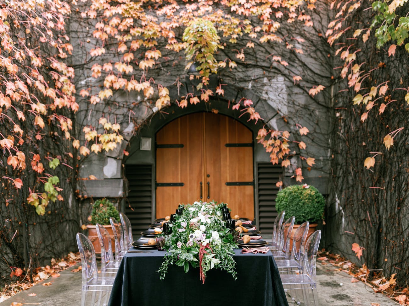 Fall tablescape with foliage table runner, black velvet linen, ghost chairs, and backdrop of ivy and fall-hued florals on stone wall | PartySlate