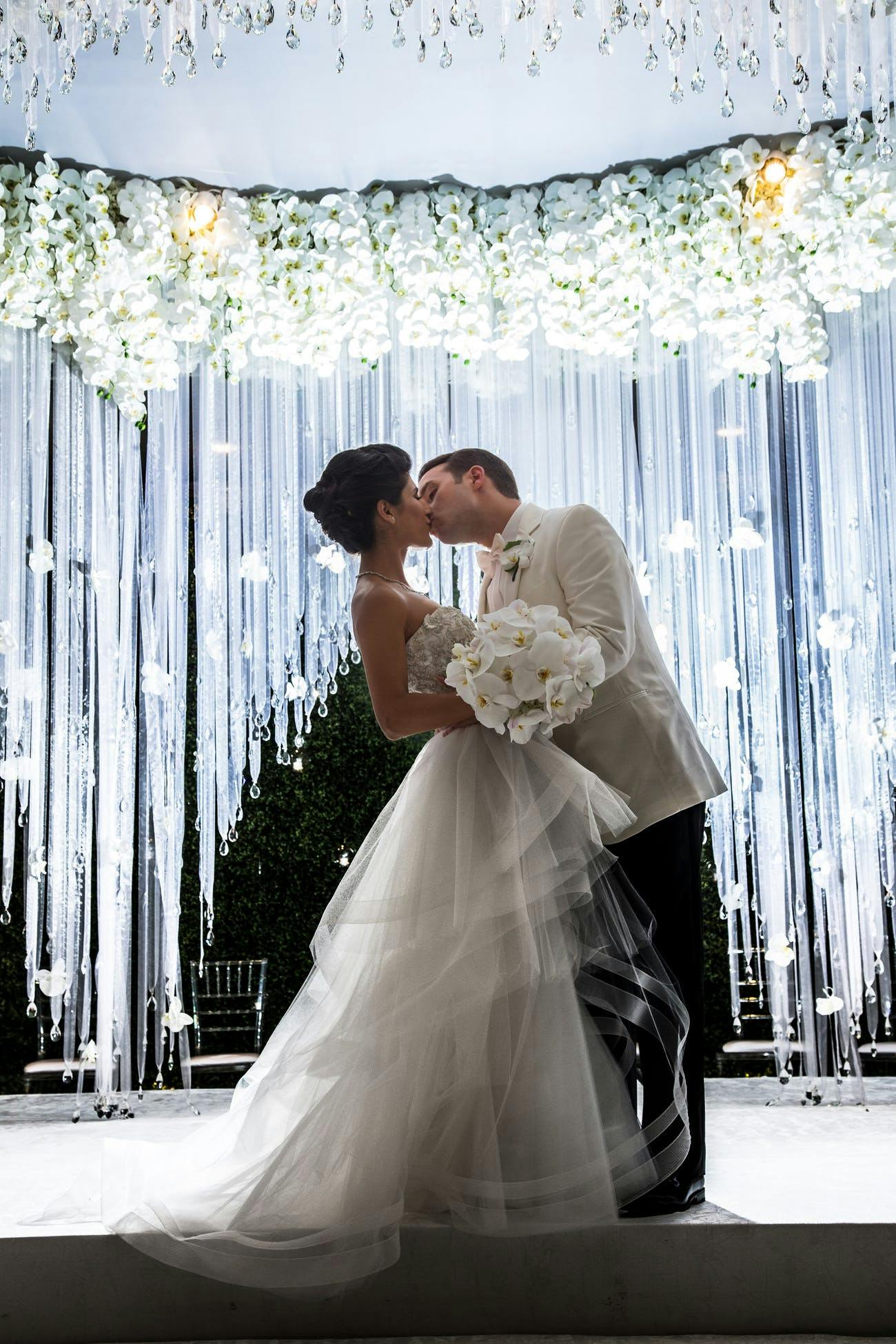 Bride and Groom Kiss in Front of Shimmering Tassel Backdrop Crowned with White Florals | PartySlate