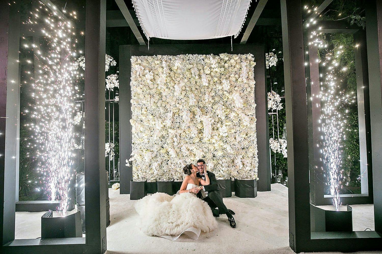 Bride and groom sit on white floor in front of black and white backdrop with cold fireworks at The Beverly Hills Hotel | PartySlate