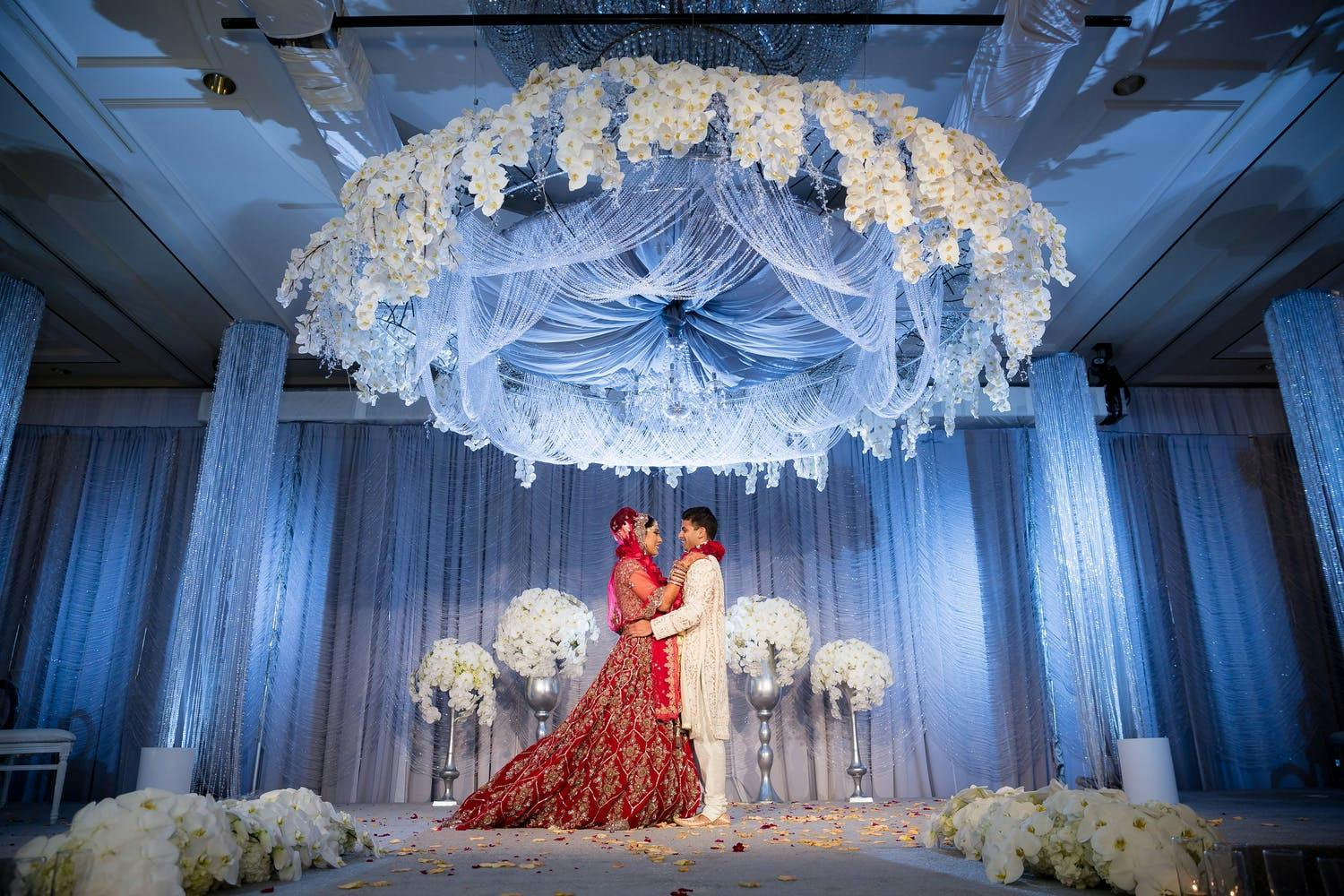Bride and groom underneath ethereal ceiling installation of shimmering tassels and white orchids and blue uplighting at Indian Wedding | PartySlate