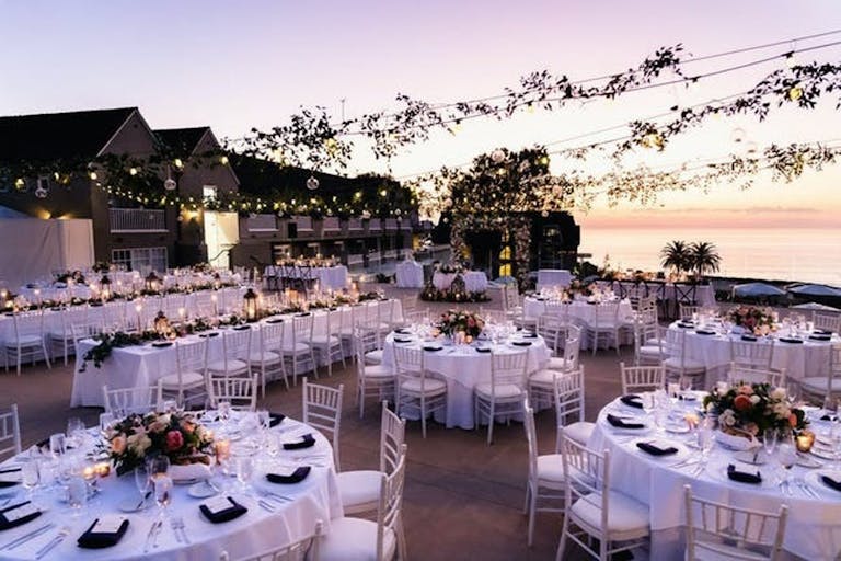 Romantic Wedding Reception at Sunset With Greenery On String Lights Draped Above Tables | PartySlate