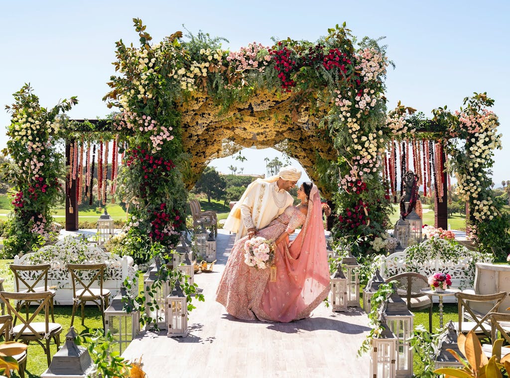 Groom dips bride in pink dress beneath lavish mandap decorated with organic greenery and pink and red florals | Partyslate