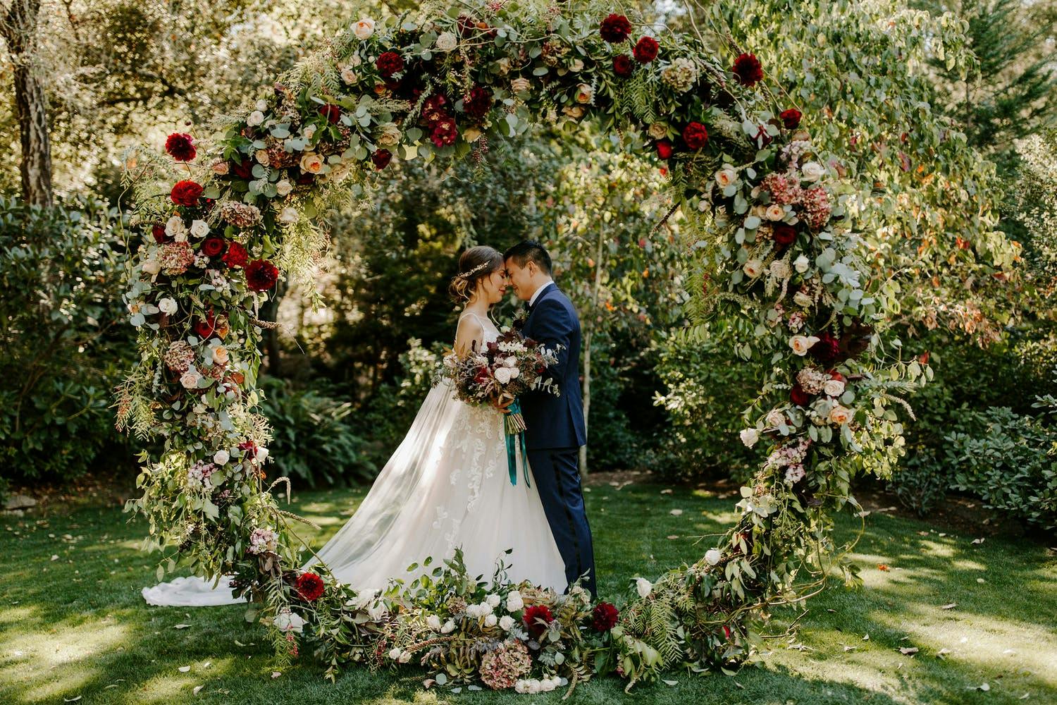 Bride and groom stand behind circular wedding arch of lavish greenery and red floral details | PartySlate