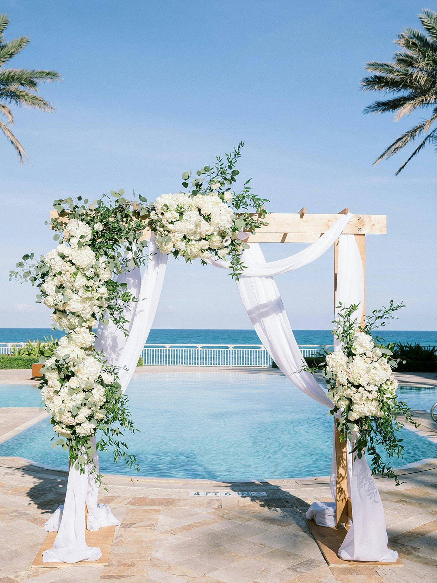Outdoor wooden wedding arch with white drapery and white flowers and greenery with pool and ocean in backdrop | PartySlate