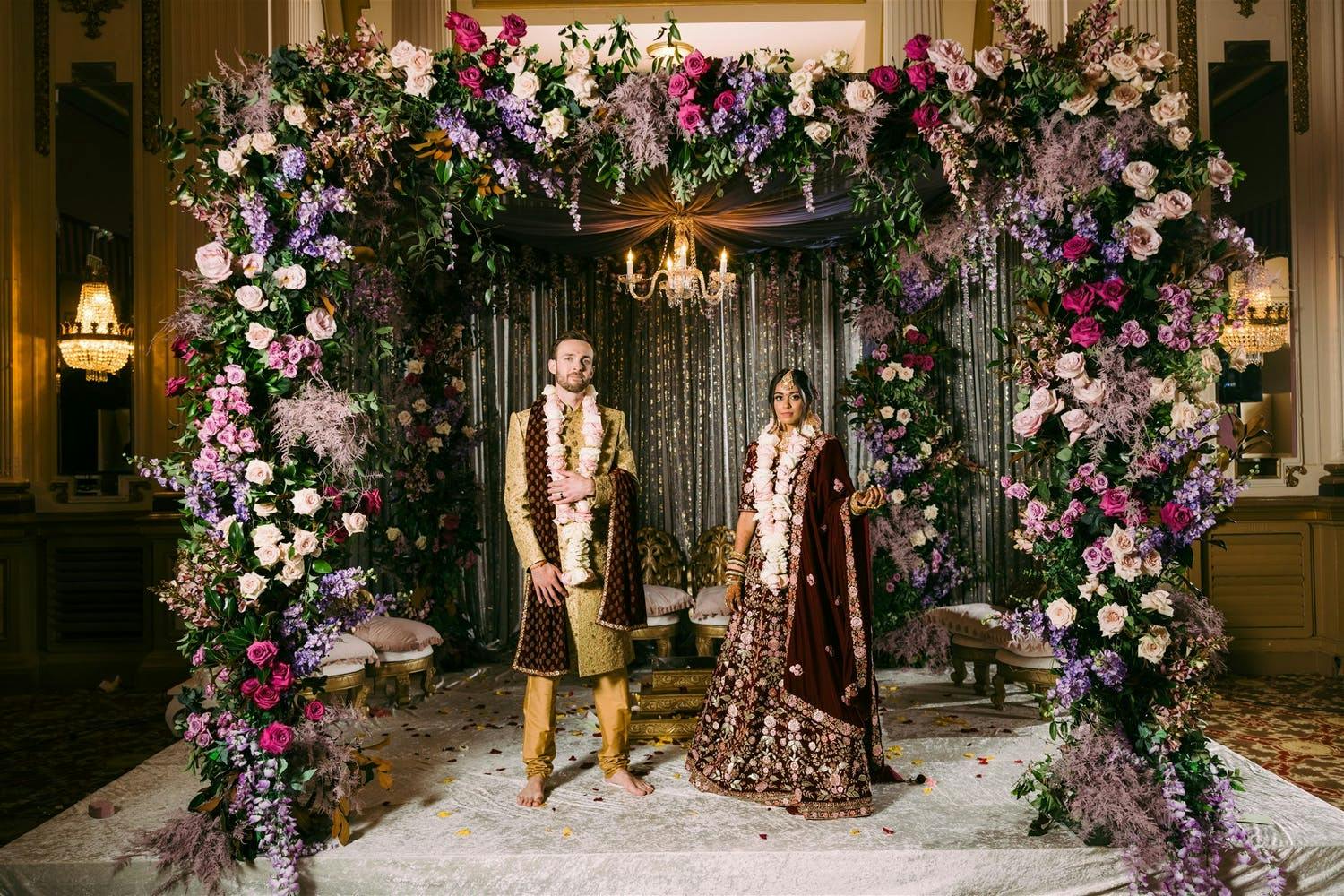 Bride and groom under lush floral mandap in pinks and purples for fusion wedding | PartySlate