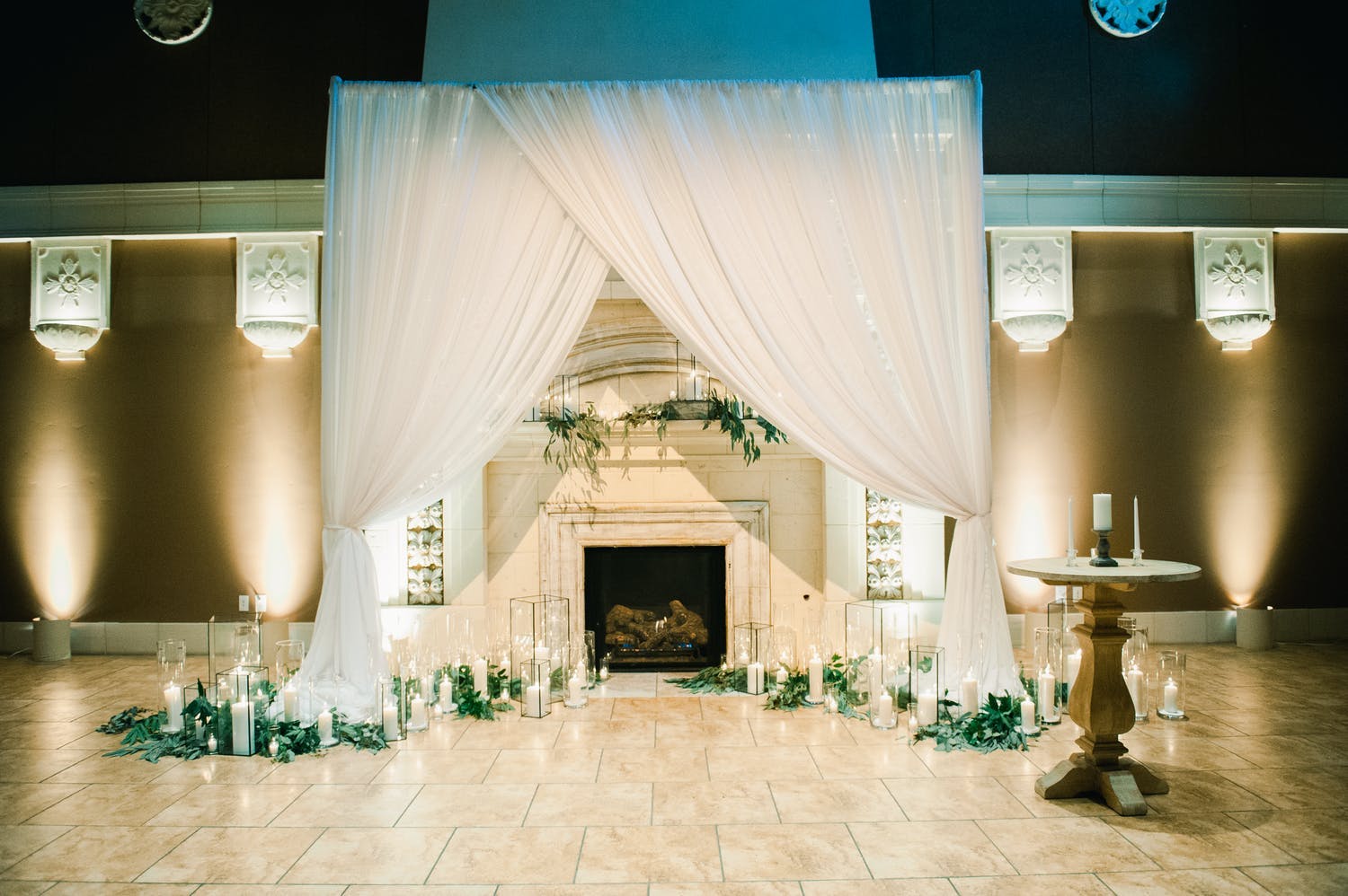 Fireplace ceremonial backdrop with white drapery, candlelight, and greenery | PartySlate