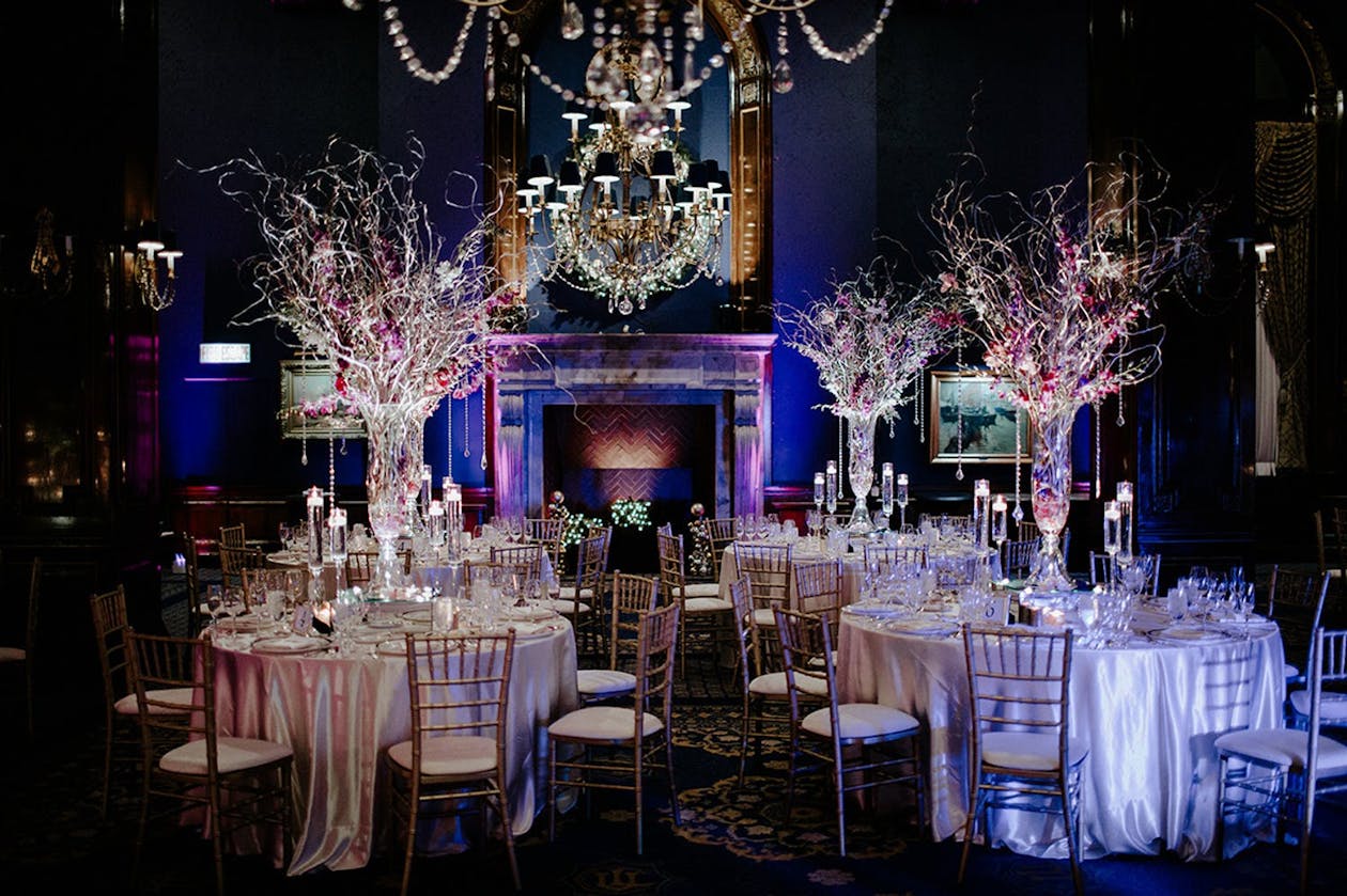 Cozy wedding venue with deep blue walls, purple uplighting, and winter wedding centerpieces of bare branches and delicate pink blooms | PartySlate