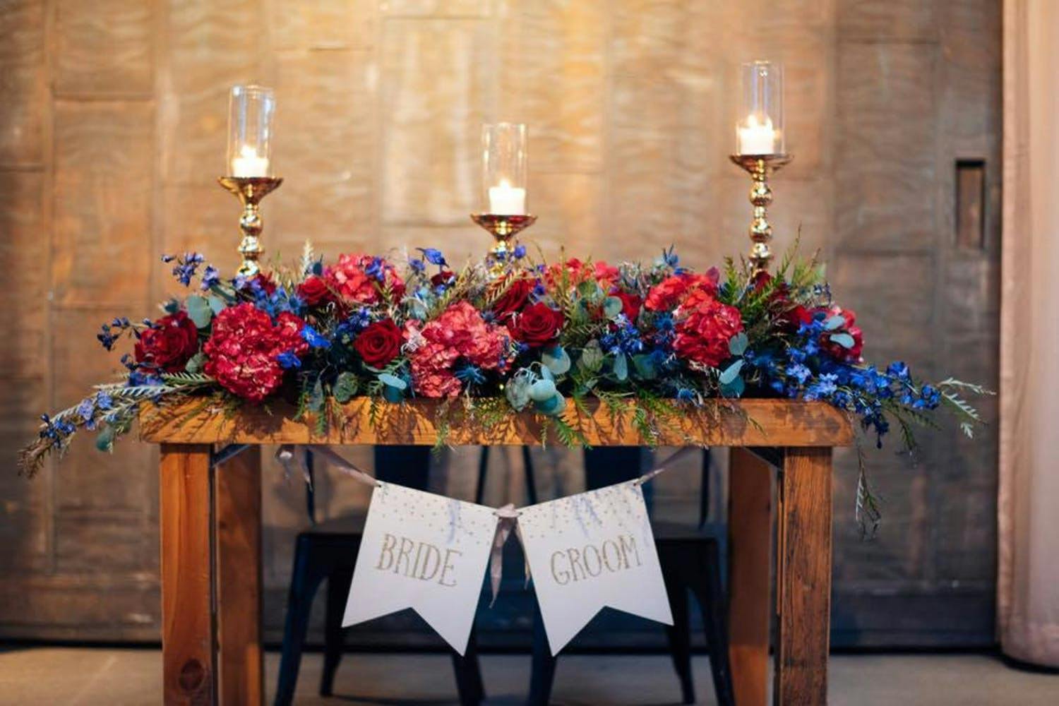 Wooden sweetheart table with red and blue flowers and golden candles | PartySlate